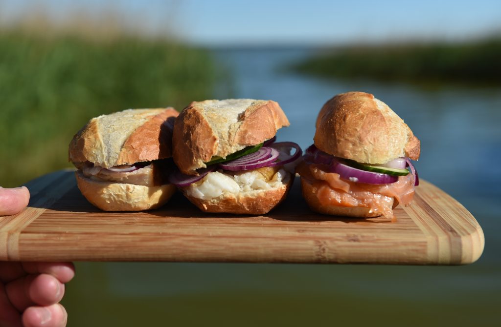 3 Fischbrötchen auf einem Brett, mit roten Zwiebeln und Gurken.